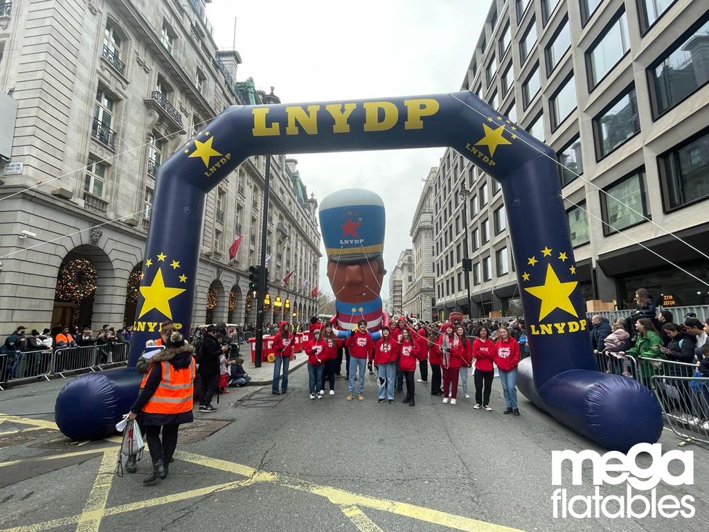 large inflatable LNYDP parade arch with stars and volunteers marching beneath a giant inflatable character balloon on a busy London street lined with spectators