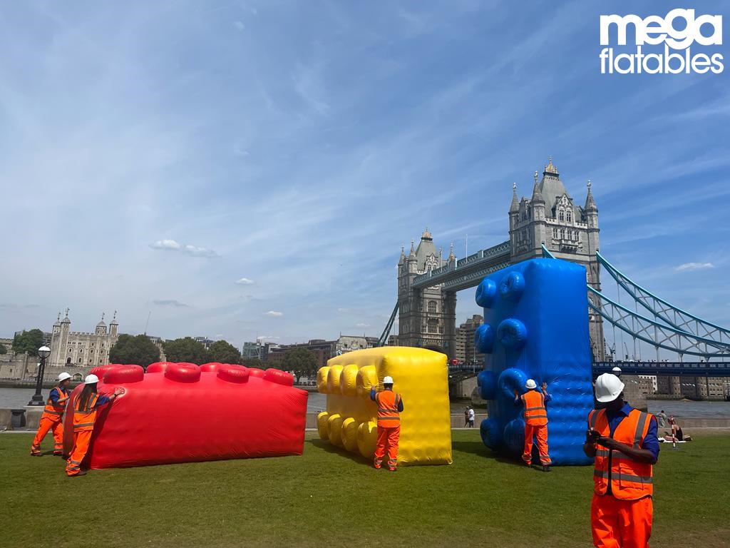 Large red, yellow, and blue inflatable blocks resembling oversized LEGO bricks are set up on a grassy area near London’s Tower Bridge. Workers in orange safety gear stand nearby, preparing the installation. Created by Mega Inflatables, this eye-catching display was designed for a Betfair promotional event.