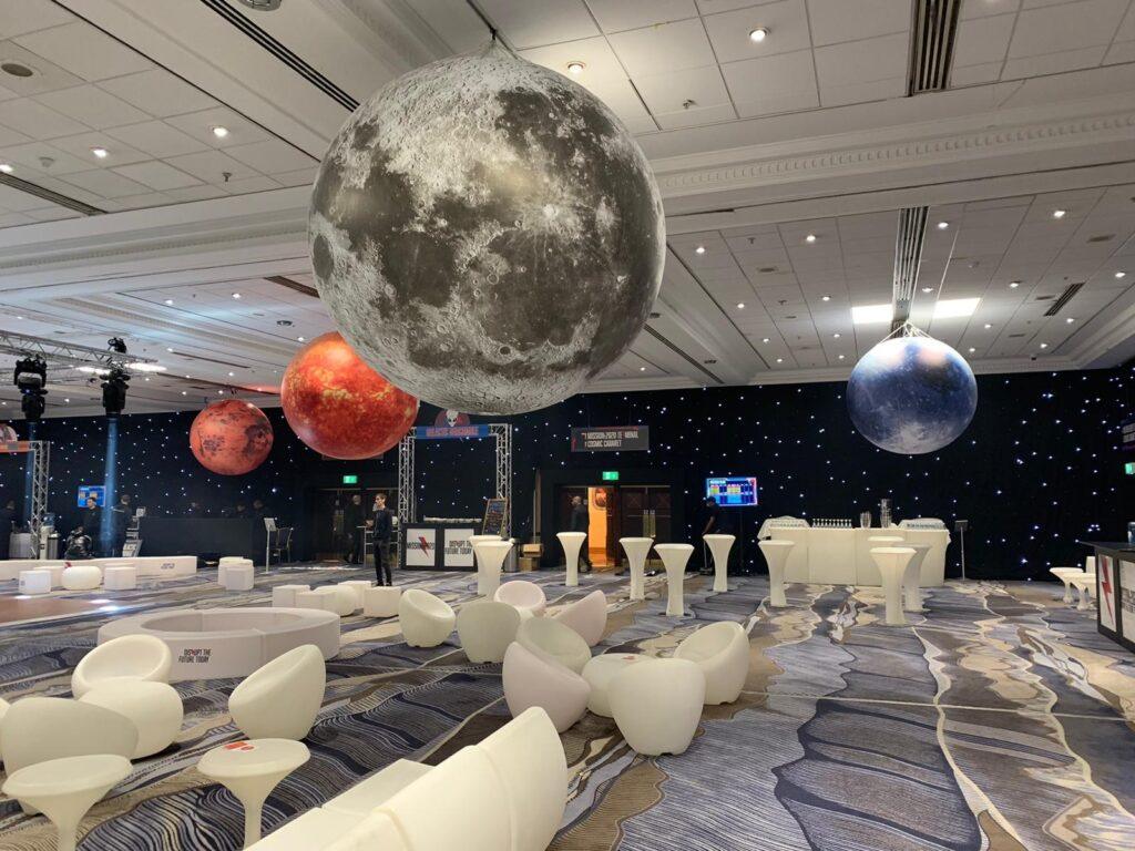 A modern event space decorated with large inflatable planets suspended from the ceiling, creating a space-themed atmosphere with ambient lighting and contemporary white furniture.