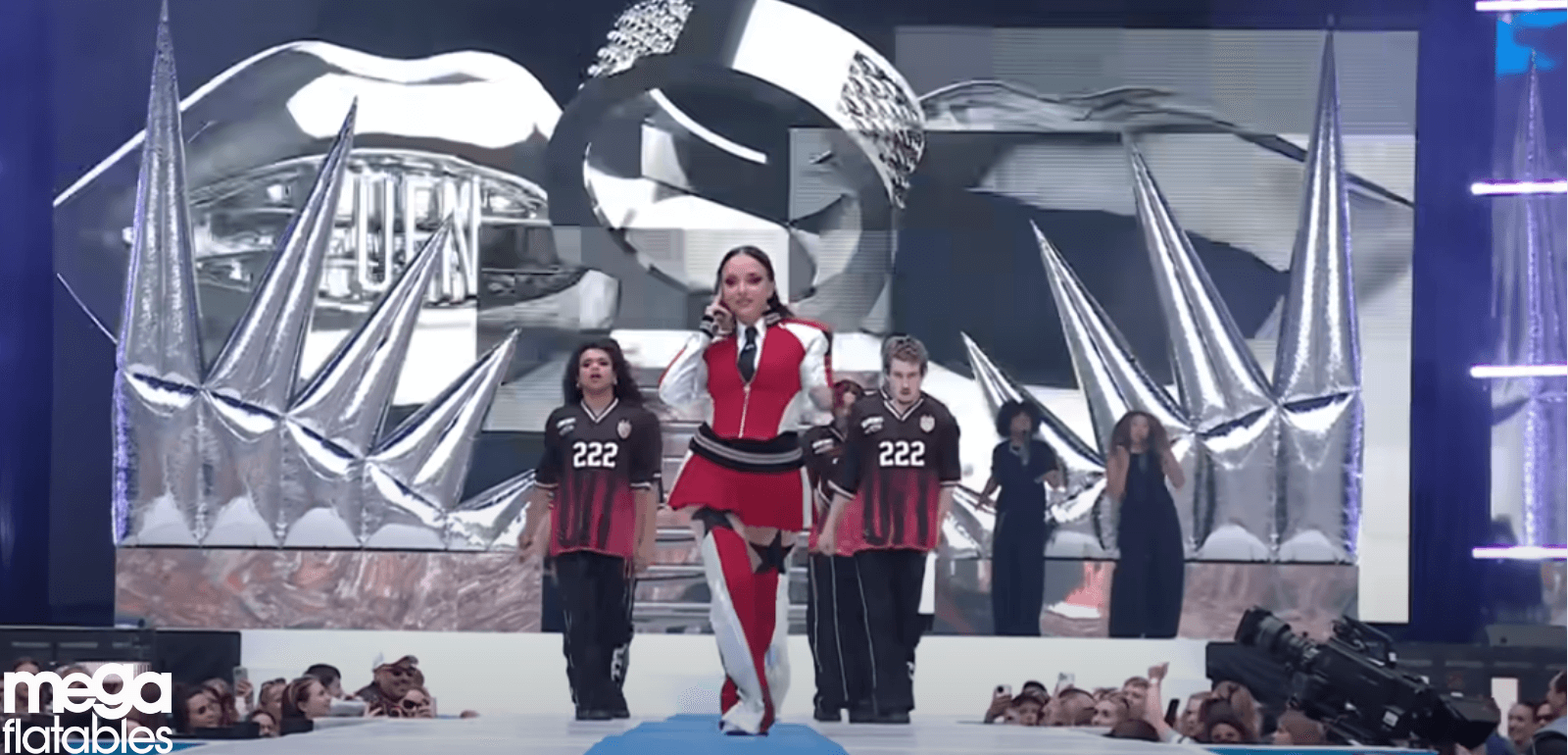 A performer sings on stage in front of large silver inflatable spike structures with dancers and a big screen backdrop showing metallic rings. The inflatables, created by Mega Inflatables, add a dramatic, futuristic element to the concert stage design.